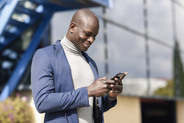 Smiling businessman wearing blue suit and grey turtleneck pullover looking at cell phone - JSMF00878