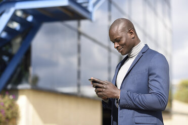Smiling businessman wearing blue suit and grey turtleneck pullover looking at cell phone - JSMF00879