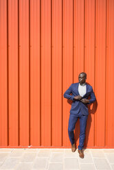 Porträt eines Geschäftsmannes in blauem Anzug und grauem Rollkragenpullover vor einer orangefarbenen Wand stehend - JSMF00897