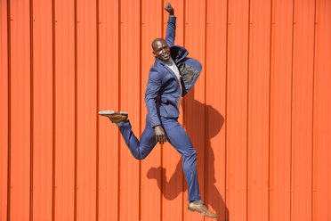 Smiling businessman jumping in the air in front of orange wall - JSMF00899