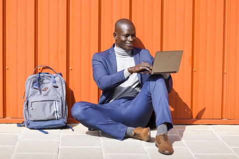 Modischer Geschäftsmann sitzt vor einer orangefarbenen Wand und benutzt einen Laptop, lizenzfreies Stockfoto