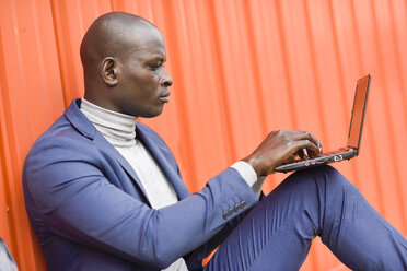 Fashionable businessman sitting in front of orange wall using laptop - JSMF00905