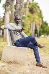 Porträt eines lächelnden Geschäftsmannes, der auf einer Bank sitzt und mit Kopfhörern und Smartphone Musik hört - JSMF00917
