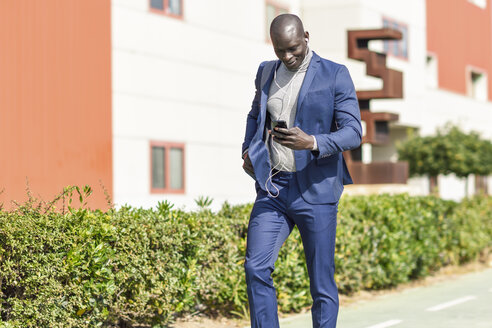 Businessman wearing blue suit listening music with earphones and smartphone - JSMF00919