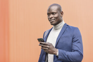 Portrait of smiling businessman with smartphone in front of orange wall - JSMF00922