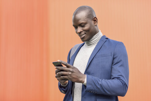 Lächelnder Geschäftsmann steht vor einer orangefarbenen Wand und schaut auf sein Smartphone - JSMF00923