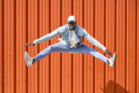 Mann in lässiger Jeanskleidung springt vor einer orangefarbenen Wand in die Luft, lizenzfreies Stockfoto