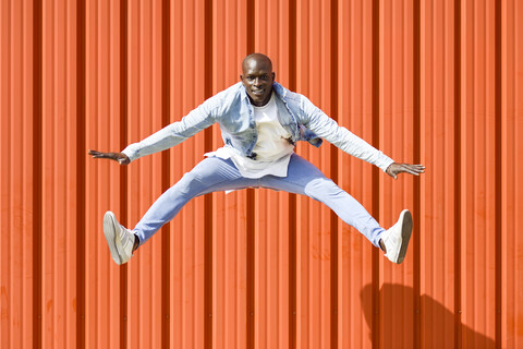 Mann in lässiger Jeanskleidung springt vor einer orangefarbenen Wand in die Luft, lizenzfreies Stockfoto