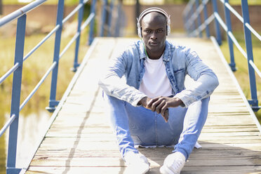 Portrait of man wearing casual denim clothes sitting on footbridge listening music with headphones - JSMF00933