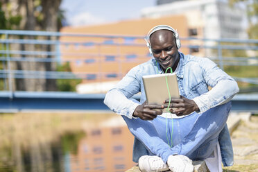 Mann in legerer Jeanskleidung hört Musik mit Kopfhörern und digitalem Tablet im Freien - JSMF00936