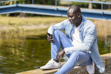 Mann in legerer Jeanskleidung sitzt in der Nähe eines Flusses und benutzt ein Smartphone - JSMF00937