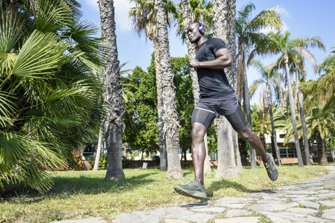 Spanien, Mann in schwarzer Sportkleidung, der in einem Park läuft und mit drahtlosen Kopfhörern Musik hört, lizenzfreies Stockfoto
