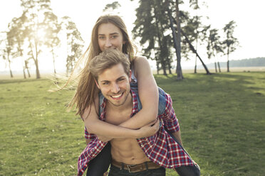 Porträt eines glücklichen jungen Mannes, der seine Freundin auf einer Wiese huckepack trägt - PNEF01334