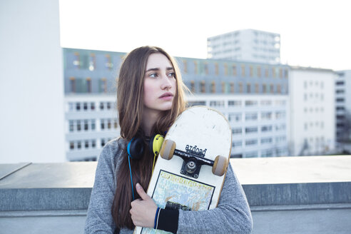 Seriöse junge Frau mit Skateboard und Kopfhörern in der Stadt, die zur Seite schaut - PNEF01354