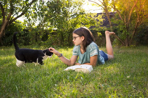 Mädchen mit Buch auf einer Wiese im Garten liegend, Katze kitzelnd, lizenzfreies Stockfoto