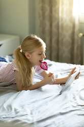 Blondes Mädchen liegt auf dem Bett und leckt einen Lutscher, während sie ein digitales Tablet benutzt - JPF00368