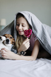 Lächelndes Mädchen auf dem Bett liegend mit Lutscher kitzelt ihren Hund - JPF00373