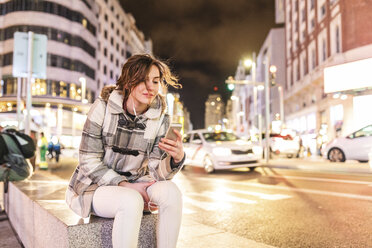 Spanien, Madrid, junge Frau in der Stadt bei Nacht, die ihr Smartphone benutzt und Kopfhörer trägt - WPEF01395