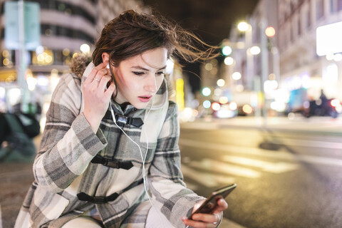 Spanien, Madrid, junge Frau in der Stadt bei Nacht, die ihr Smartphone benutzt und Kopfhörer trägt, lizenzfreies Stockfoto