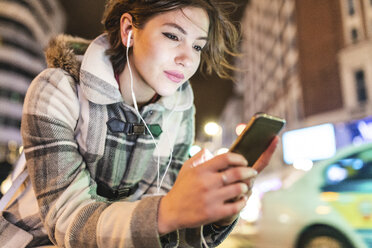 Spain, Madrid, young woman in the city at night using her smartphone and wearing earphones - WPEF01398