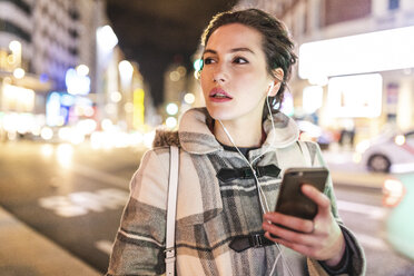 Spanien, Madrid, junge Frau in der Stadt bei Nacht, die ihr Smartphone benutzt und Kopfhörer trägt - WPEF01399