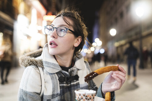 Spain, Madrid, young woman in the city at night eating typical churros with chocolate - WPEF01407