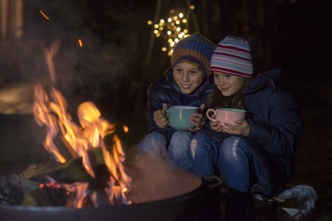 Junge und Mädchen mit Tassen sitzen abends am Feuer, lizenzfreies Stockfoto