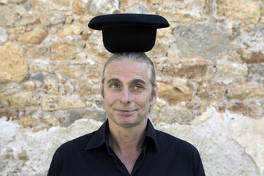 Portrait of man at stone wall balancing a bowler hat on his head - PSTF00350