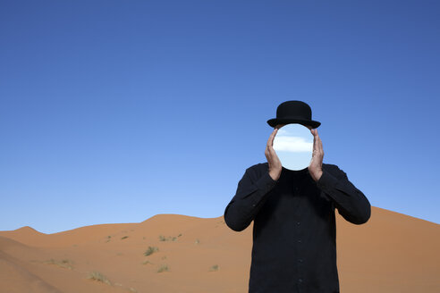 Marokko, Merzouga, Erg Chebbi, Mann mit Melone, der sich in einer Wüstendüne einen Spiegel vor das Gesicht hält - PSTF00394