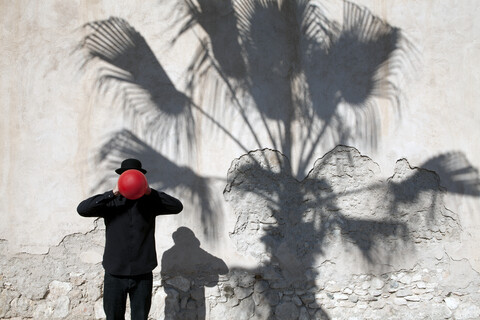 Marokko, Essaouira, Mann mit Melone, der einen roten Luftballon vor sein Gesicht hält, an einer Mauer, lizenzfreies Stockfoto