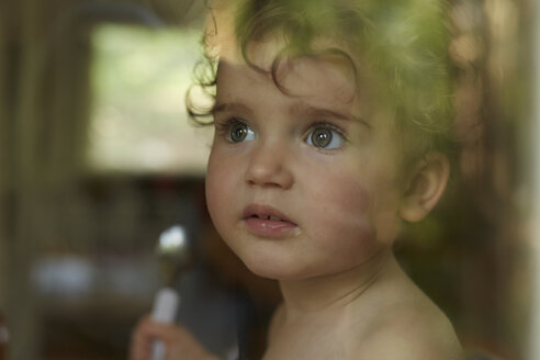 Portrait of little girl behind windowpane eating - AMEF00048