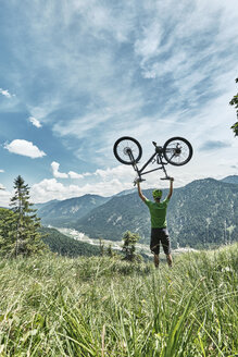 Deutschland, Bayern, Isartal, Karwendelgebirge, Mountainbiker bei einem Ausflug, der sein Fahrrad auf einer Almwiese hochhebt - WFF00076