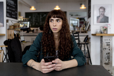 Junge Frau mit Mobiltelefon in einem Café, die sich umschaut - FLLF00069