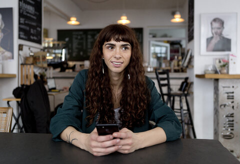 Lächelnde junge Frau mit Handy in einem Cafe, die sich umschaut - FLLF00070