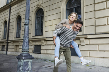 Happy man giving woman piggyback ride on pavement in the city - PNEF01425