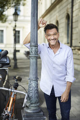 Porträt eines lächelnden Geschäftsmannes mit Fahrrad, der sich gegen einen Laternenpfahl in der Stadt lehnt - PNEF01438