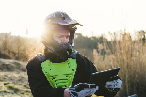 Porträt eines Motocross-Fahrers mit Blick auf ein Tablet bei Sonnenuntergang - FBAF00339