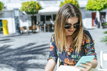 Spanien, Cadiz, Vejer de la Frontera, junge Frau sitzt in einem Straßencafé und schaut auf ihr Mobiltelefon - KIJF02472