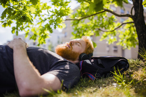 Gelassener Mann mit Bart, der in einem sonnigen Park mit Kopfhörern Musik hört - FSIF03845