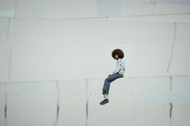 Porträt selbstbewusster junger Mann mit Afro, der auf einer sonnigen Stadtmauer sitzt - FSIF03866