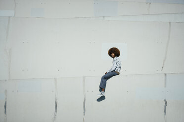 Portrait confident young man with afro sitting on sunny urban wall - FSIF03866