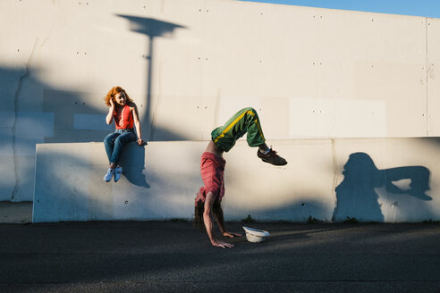 Young woman watching man do handstand along urban wall - FSIF03924