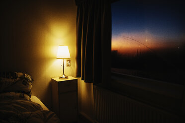 Bedside lamp illuminating bedroom corner next to window with view of dusk sky - FSIF03933