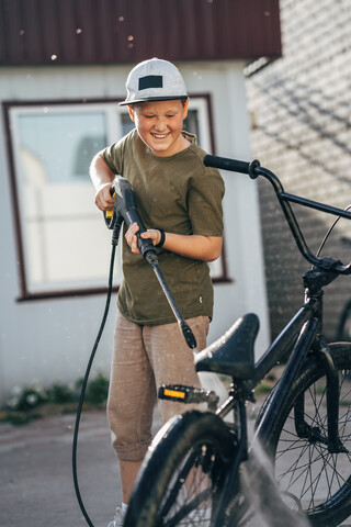 Junge wäscht BMX-Fahrrad mit Hochdruckreiniger auf dem Hof, lizenzfreies Stockfoto