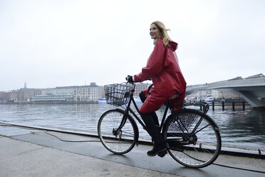 Denmark, Copenhagen, happy woman riding bicycle at the waterfront in rainy weather - ECPF00660