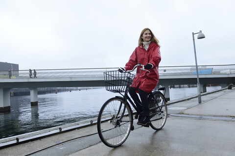 Dänemark, Kopenhagen, glückliche Frau beim Fahrradfahren am Wasser bei regnerischem Wetter, lizenzfreies Stockfoto