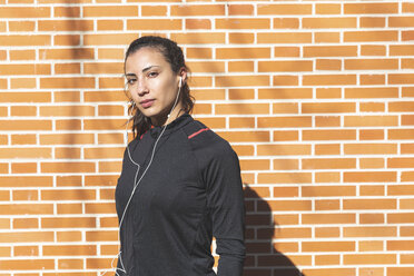 Porträt einer sportlichen jungen Frau mit Kopfhörern an einer Backsteinmauer - WPEF01432