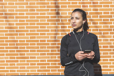 Portrait of sporty young woman with earphones and cell phone at a brick wall - WPEF01433