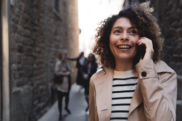 Lächelnde Frau, die in einer Gasse mit einem Handy telefoniert - FMOF00490