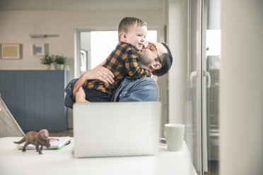 Vater sitzt am Tisch im Home Office und umarmt seinen Sohn - UUF16864